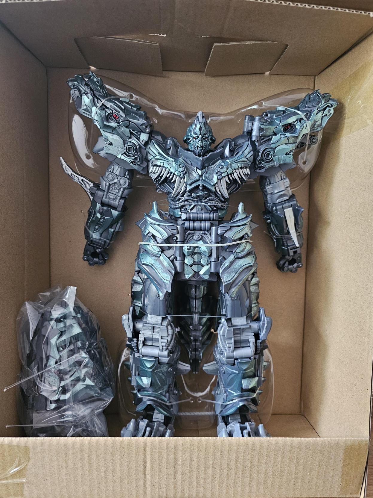A picture of the Grimlock figure sitting in the inner cardboard tray. Next to him is a small bag of parts to complete the figure